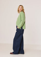 A woman stands in profile wearing a light sage green fuzzy knit sweater, dark blue wide-leg jeans, and brown shoes against a plain off-white studio background.