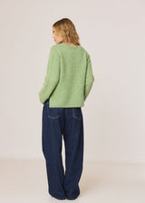 A woman is shown from behind wearing a light green textured knit sweater and dark blue wide-leg jeans, standing against a plain off-white studio background.