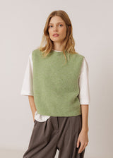 A woman modeling a light sage green knit sleeveless vest layered over a white short-sleeve top and dark brown wide-leg trousers, set against a plain light gray studio background.