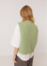 A woman viewed from behind wearing a light sage green knit vest over a white short-sleeve top and dark brown pants, set against a plain off-white studio background.