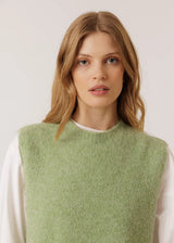 A woman with blonde hair wearing a light sage green knit sweater vest over a white long-sleeve shirt, photographed against a plain light gray studio background.