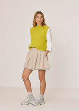 A woman modeling a bright lime green knit sweater vest over a white long-sleeve shirt, paired with a beige pleated linen skirt, gray socks, and silver sneakers, against a plain off-white studio background.