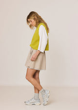 A woman modeling a bright yellow-green knit vest over a white long-sleeve shirt, paired with a beige pleated mini skirt, gray socks, and light gray sneakers, set against a plain beige studio background.