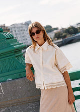 A woman wearing a cream short-sleeve lace-trim button-up shirt and a beige skirt, leaning against a green bridge railing with a river and city buildings in the background.