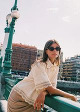 A woman wearing a cream lace short-sleeve blouse and beige skirt leans on a green bridge railing in an outdoor city setting.