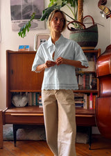 A woman stands in a cozy room wearing a light blue short-sleeve button-up shirt with lace trim and beige wide-leg pants, positioned in front of a wooden bookcase and piano.