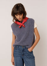 A model wearing a sleeveless, textured slate blue knit vest over a white t-shirt, styled with a red ruffled neck scarf and blue jeans, against a plain light background.