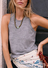 A woman wearing a gray ribbed knit sleeveless tank top, styled with a patterned cream scarf and a beaded necklace, leaning against a dark wooden door.