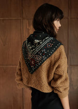 A woman shown from the back wearing a brown cable-knit cropped sweater and a black patterned bandana draped over her shoulders, set against a dark wood-paneled background.