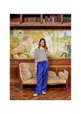 A woman stands in a vintage room with a large classical painting on the wall, wearing a black and white striped long-sleeve top and vibrant royal blue wide-leg trousers.