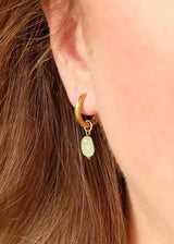 A close-up of a person's ear wearing a gold crescent-shaped huggie hoop earring with a dangling light green teardrop stone.