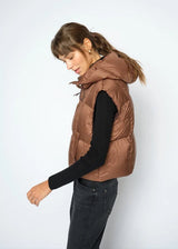 A woman wearing a brown puffer vest over a black long-sleeve top and dark jeans, standing against a light gray studio background.
