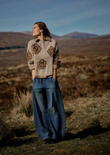 Woman standing in a field with mountains in the background wearing a patterned sweater and jeans.