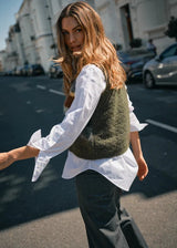 Woman in a white shirt and green vest standing on a street.