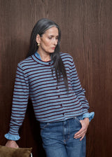 A woman wearing a blue and navy striped knit cardigan with ruffled cuffs and a button front, paired with blue jeans, standing against a dark wood-paneled wall.