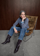 A woman with gray hair sits in a brown upholstered chair against a dark wood-paneled wall, wearing a blue and gray striped long-sleeve top, blue jeans, and dark pointed-toe boots.