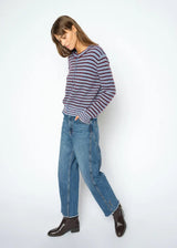 A woman modeling a long-sleeve striped knit top with blue jeans and dark brown leather ankle boots, standing against a plain white studio background.