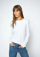 A woman wearing a plain white long-sleeve crewneck top and blue jeans, standing against a light gray studio background.