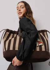 A woman wearing a black long-sleeve top carries a large brown and beige striped canvas tote bag with dark brown drawstring handles against a plain light gray background.