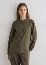 A woman modeling a long-sleeve button-front shirt in olive green with a band collar, paired with brown pants, against a plain light gray wall.