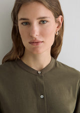 A close-up portrait of a woman wearing a muted olive-brown button-front top with a rounded collar and small silver hoop earrings against a plain light gray background.