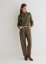 A woman stands against a plain light gray studio background wearing an olive green long-sleeve button-up shirt with a cowl neck, brown wide-leg pants, a patterned belt, and brown heeled ankle boots.