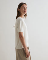 A woman in a side profile wearing a short-sleeve cream-colored blouse and beige trousers, standing against a plain light gray studio background.