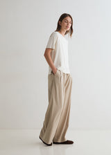 A woman stands in profile wearing a loose white short-sleeve top and wide-leg beige trousers against a clean white studio background.