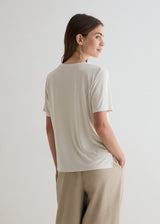 A woman shown from the back wearing a short-sleeve cream-colored t-shirt and beige trousers, standing against a plain light gray studio background.