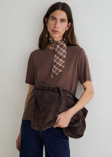 A woman models a brown short-sleeve t-shirt with a unique keyhole cutout, styled with a brown and beige plaid neck scarf and holding a matching brown suede-like shoulder bag. She is wearing dark blue jeans against a plain light gray background.