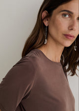 A close-up of a woman wearing long silver dangle earrings with small clear stones, paired with a brown short-sleeve top against a light gray background.