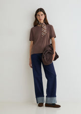 A woman stands against a plain white studio wall wearing a brown short-sleeve top, navy wide-leg jeans with cuffed hems, brown shoes, and holding a dark brown suede-like bag. She is also wearing a patterned neck scarf.