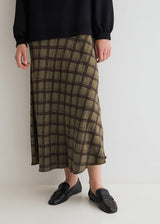 A person wearing a long olive green plaid midi skirt with a black top and black loafers, standing against a plain off-white studio background.