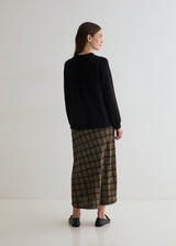 A woman is shown from the back wearing a black long-sleeve top and a brown and black plaid midi skirt, paired with black flat shoes, against a plain white studio background.