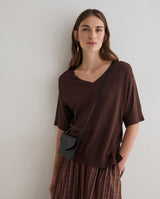 A woman wearing a dark brown short-sleeve V-neck top and a brown plaid skirt, carrying a small black crossbody bag, standing against a plain light gray wall.