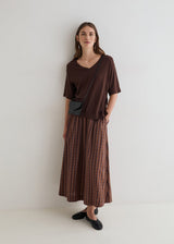 A woman stands against a plain white wall wearing a loose brown short-sleeve top, a brown and rust plaid maxi skirt, and black flats, with a small black crossbody bag.