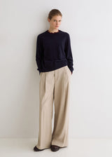 A woman stands against a plain white wall wearing a dark navy long-sleeve top with a scalloped neckline and beige wide-leg trousers.