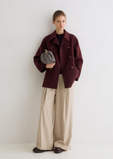 A woman stands against a plain light gray wall wearing a dark burgundy double-breasted coat over a navy top, paired with wide-leg beige trousers and dark brown shoes. She holds a gray textured clutch.