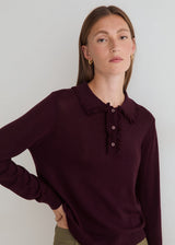 A woman wearing a long-sleeve knit polo-style sweater in deep burgundy with a ruffled collar and a three-button henley neckline, paired with olive green pants against a plain light gray studio background.