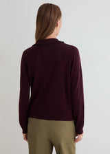 A woman viewed from the back wearing a dark burgundy knit sweater and olive green trousers, set against a plain light gray studio background.