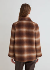 A woman viewed from the back wearing a brown and beige plaid wool-blend jacket with a high collar, paired with dark brown trousers, against a plain light gray studio background.