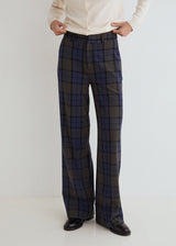 A person wearing high-waisted, wide-leg plaid trousers in dark gray and navy blue, styled with a cream button-up shirt and black leather shoes against a plain white studio background.