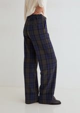 A person shown from the back wearing wide-leg plaid trousers in dark navy, gray, and black, paired with a cream top and black shoes, against a clean white studio background.