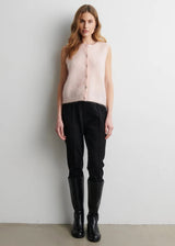A woman stands against a plain light gray wall wearing a sleeveless pale blush pink fuzzy knit vest, black pants, and black knee-high boots.
