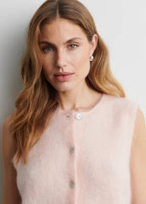 A woman with long wavy light brown hair wearing a pale pink sleeveless fuzzy knit vest with silver buttons, photographed against a plain light gray background.
