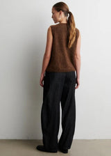 Woman wearing a brown sleeveless top and black pants against a white wall.