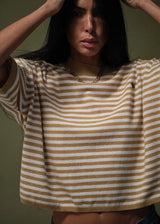 A woman wearing a cropped short-sleeve knit top with horizontal tan and off-white stripes, featuring a small dark embroidered polo player logo on the left chest, set against a dark olive background.