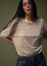 A woman wearing a beige and brown striped knit top with blue jeans, standing against a muted olive green background.