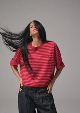 A woman with long, wind-blown black hair poses in a red and pink horizontally striped short-sleeve t-shirt and dark denim jeans against a solid gray studio background.