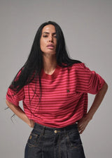 A woman modeling a red and dark pink striped short-sleeve t-shirt with a small embroidered logo on the left chest, paired with dark jeans, against a solid gray studio background.